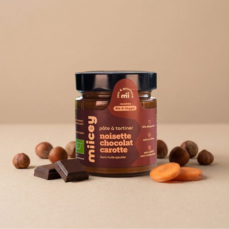 Pot de pâte à tartiner bio MIICEY Noisette, chocolat et carotte, entouré de noisettes, de carrés de chocolat noir et de rondelles de carotte.